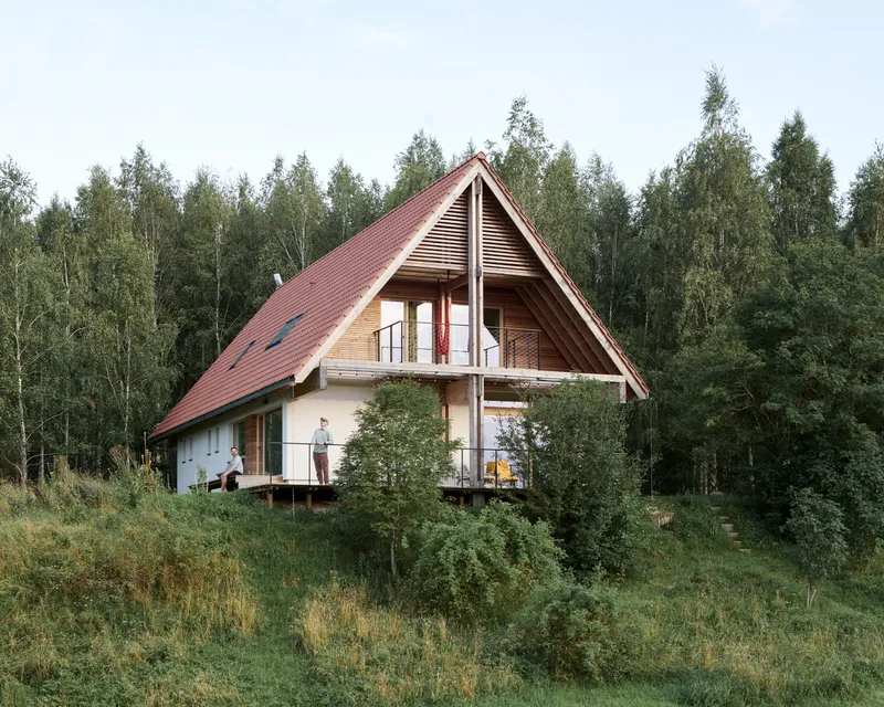 Steeply pitched red metal roof with wood cladding and balcony visible against a dense tree backdrop