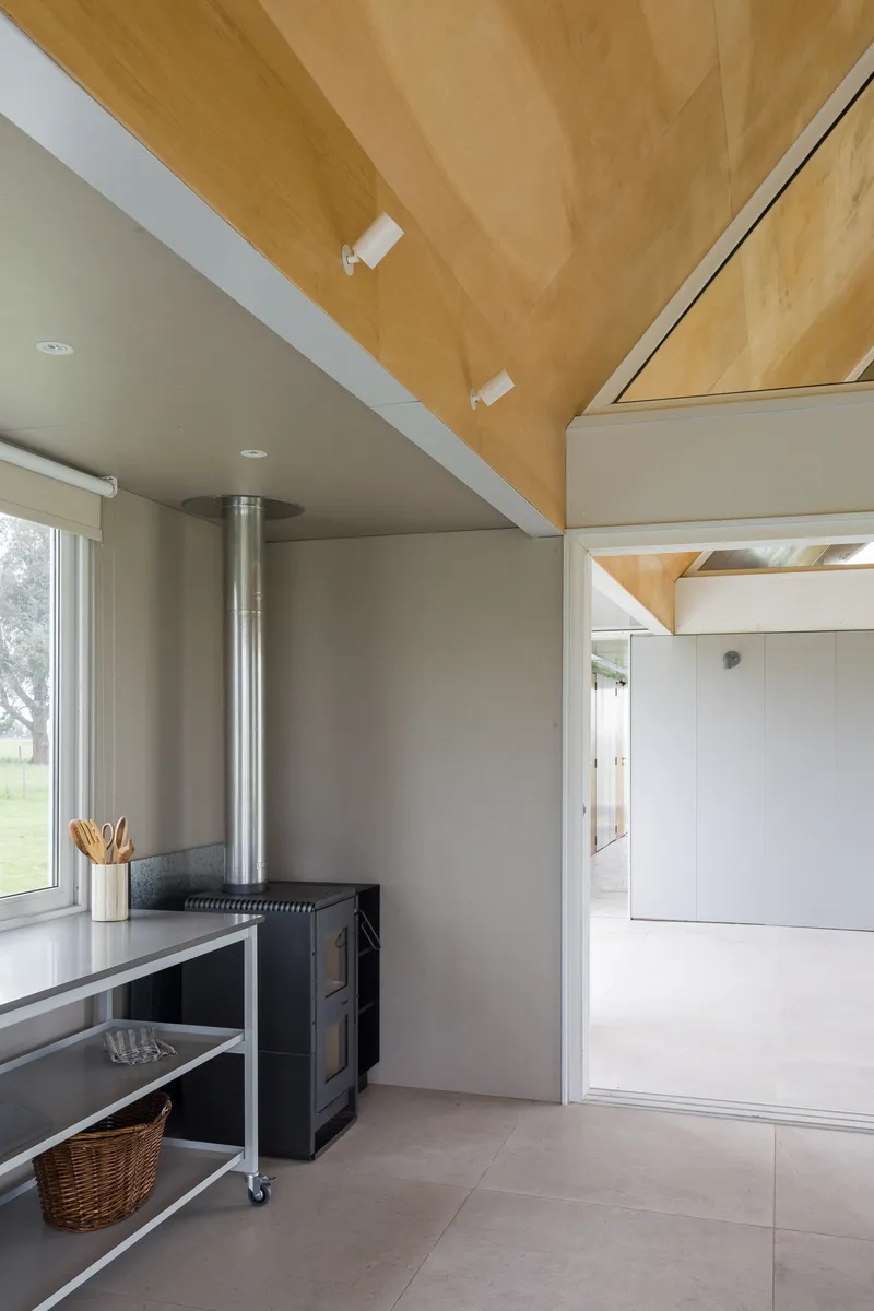 Kitchen corner with freestanding wood stove and steel shelving unit below plywood vaulted ceiling