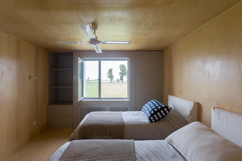 Plywood-lined bedroom with two beds and a window framing a view of distant trees and lawn