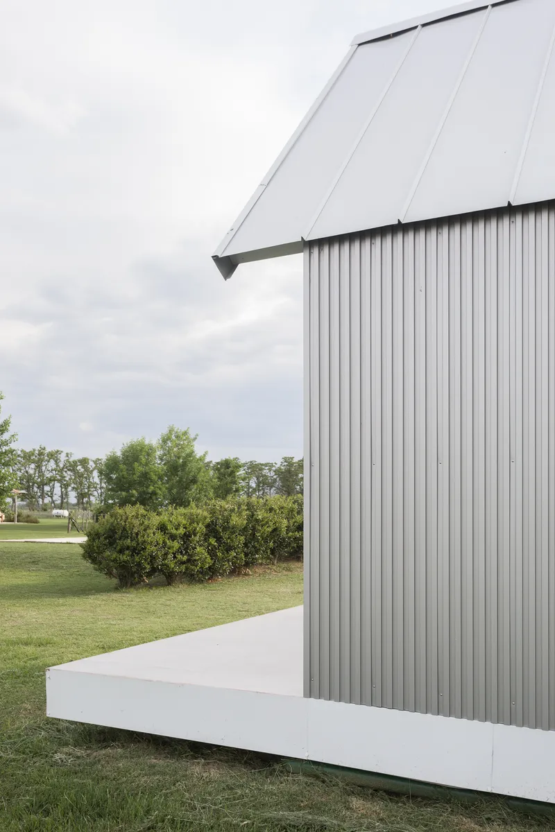 Corrugated metal volume with white plinth base and angled roof plane against a cloudy sky