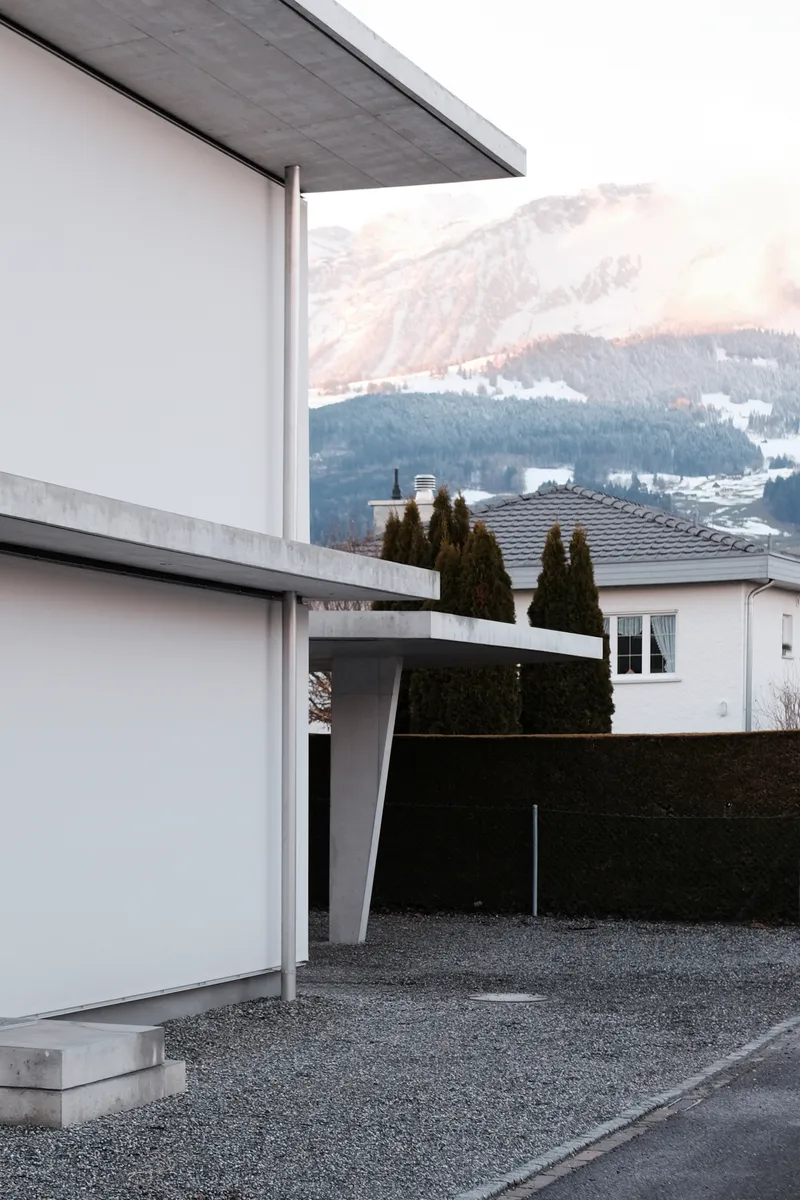 Concrete canopy supported by a slender column over a gravel drive with snowy peaks in the background