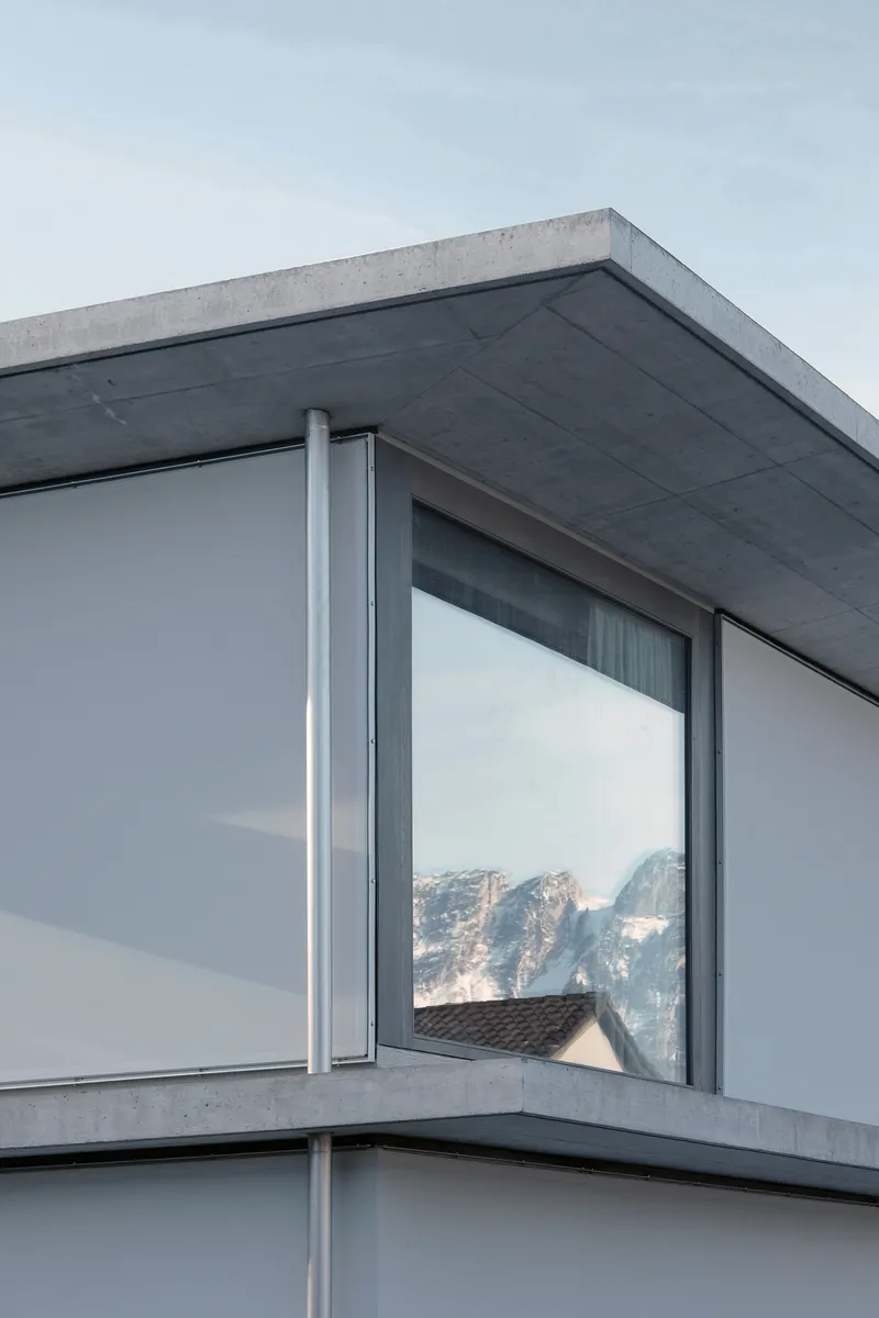 Corner detail of the concrete roof edge and floor-to-ceiling glazing reflecting misty mountain slopes
