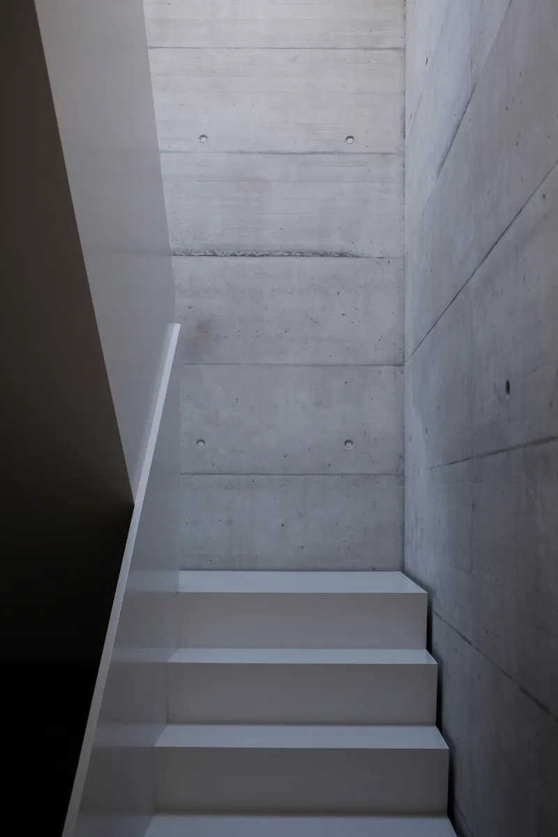 White plastered staircase ascending alongside board-formed concrete walls with visible tie holes