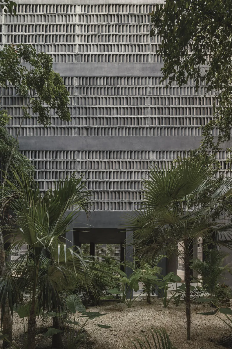 Frontal view of the repeating concrete block screen facade above the passage with palms and gravel below