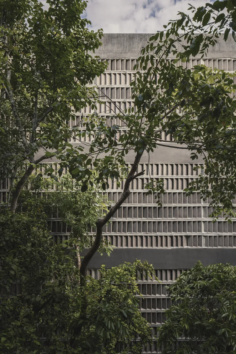 Concrete facade with horizontal brise-soleil bands framed by foreground trees in dappled sunlight
