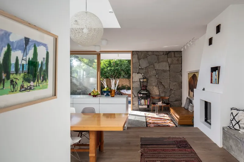 Dining area with timber table looking toward stone fireplace wall and glazed opening framing poolside tree