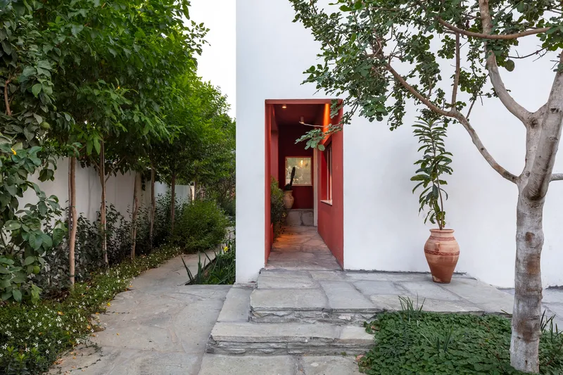 White stucco courtyard with red-framed doorway flanked by planted beds and terracotta pot