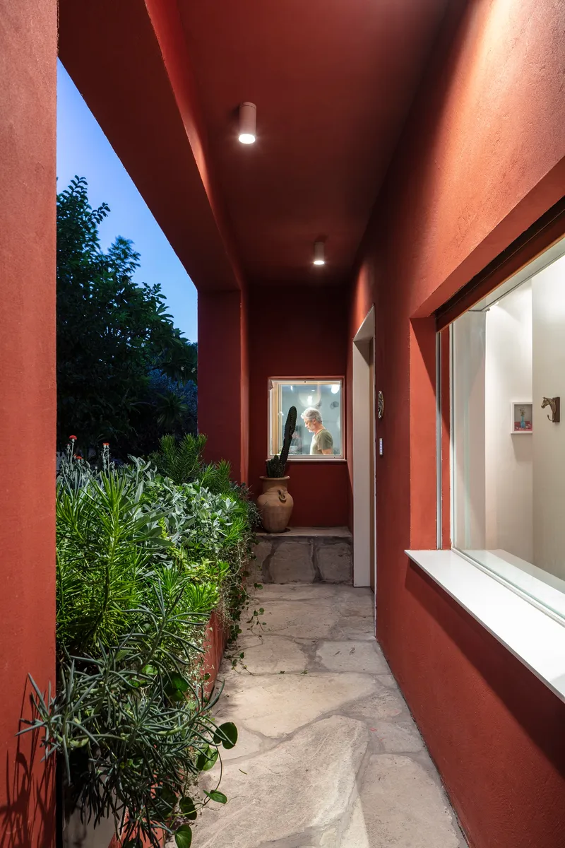 Red-painted covered walkway with recessed lighting and potted plants along stone paving at dusk