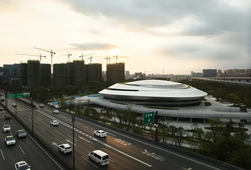 Layered elliptical roof rising above a multi-lane roadway with construction cranes in the distance