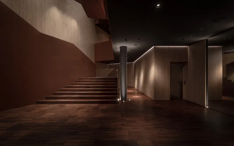Cantilevered timber staircase rising from a wood-paneled lobby with recessed linear lighting and dark ceiling