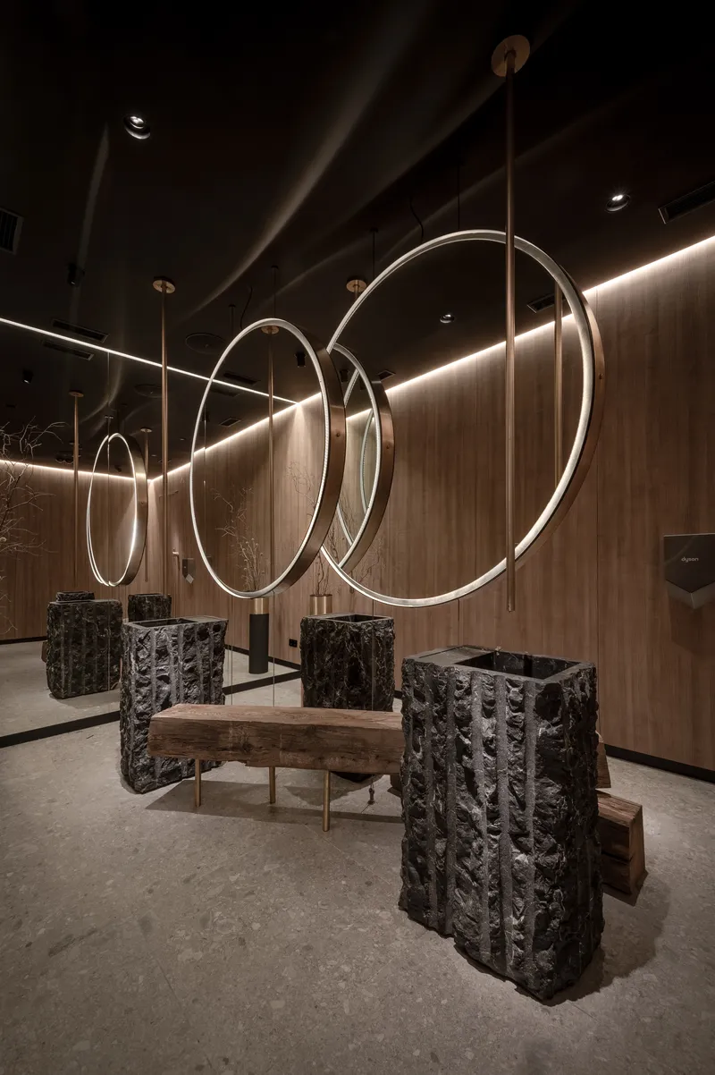 Lounge with circular metal light rings suspended above textured black stone seating blocks and timber bench