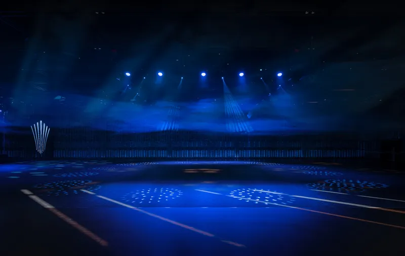 Interior arena space with blue stage lighting and perforated ceiling visible through atmospheric haze
