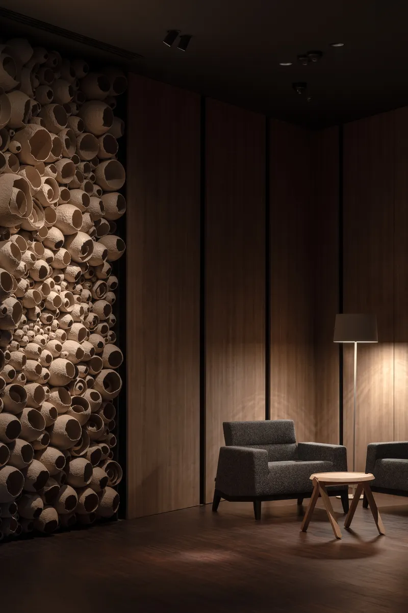 Corner view of timber cylinder wall sculpture beside wood-paneled wall with armchair and floor lamp
