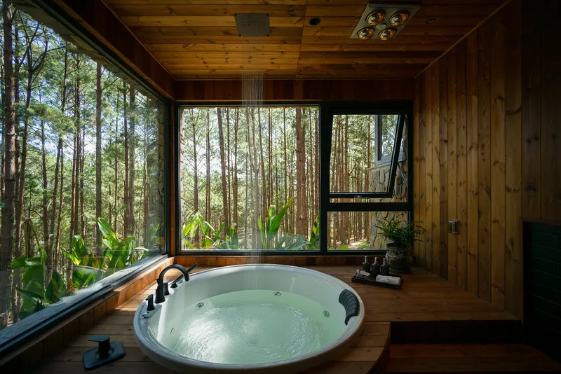 Circular jacuzzi set into timber deck framed by corner windows facing dense forest vegetation