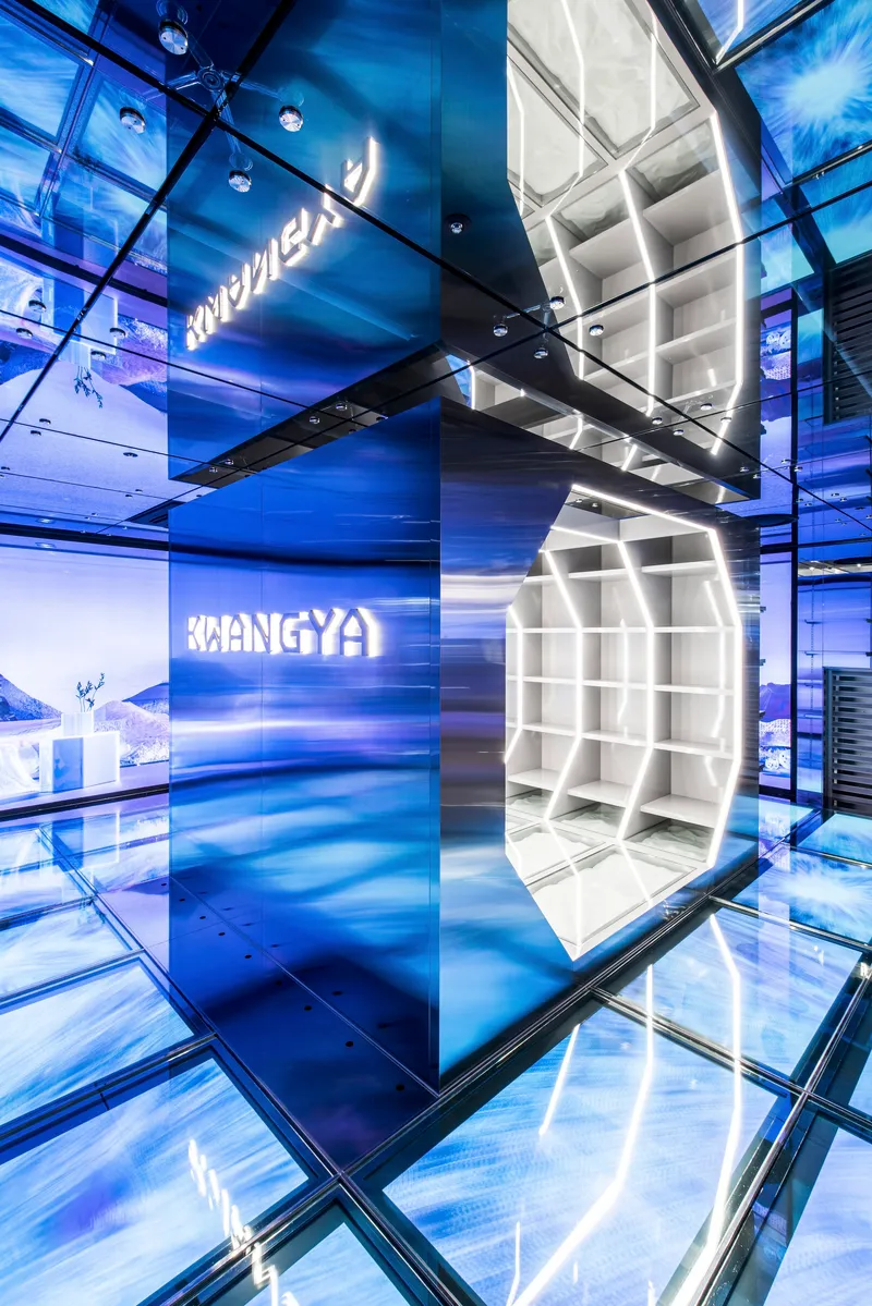 Multi-story retail atrium with illuminated blue glass walls and suspended white display cube under glass floor