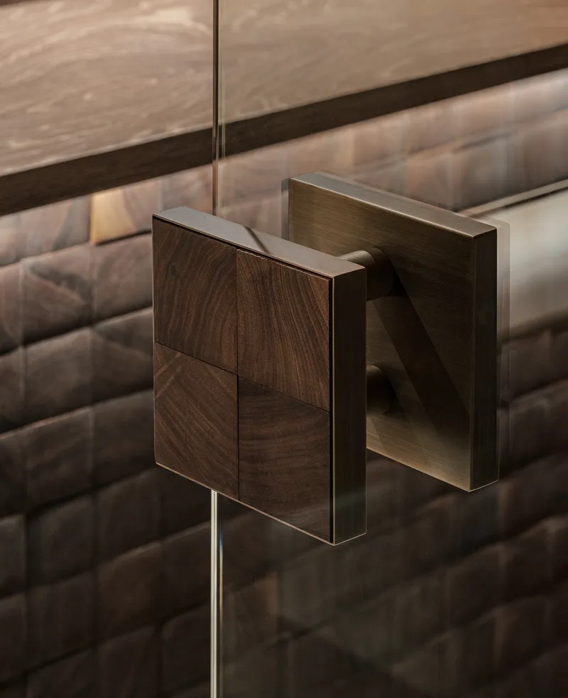 Close-up of a brushed metal door handle with walnut veneer plate against textured tile wall