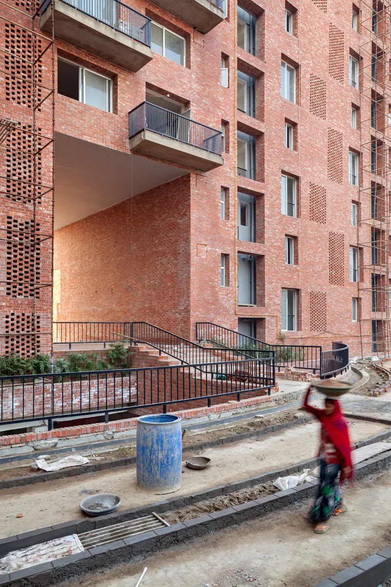 Red brick facade with perforated screens and recessed balconies, with a cyclist in motion on the construction site below