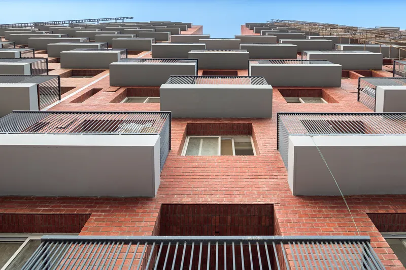 Upward view of the terraced facade showing alternating brick volumes and corrugated metal sunshades under a clear sky