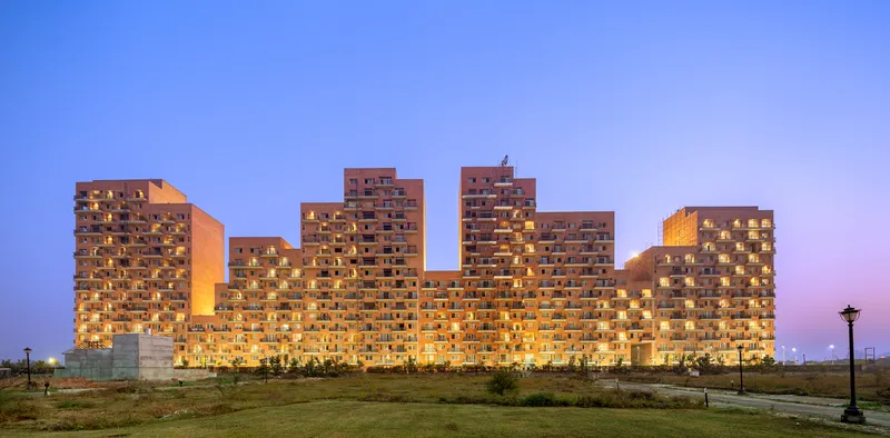 Stepped residential block with illuminated balconies and perforated brick screens photographed at twilight from an open field