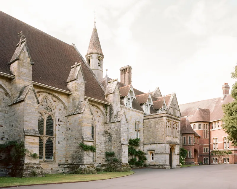 Stone chapel with pitched roof and bell tower adjoining pitched-roof buildings under overcast sky