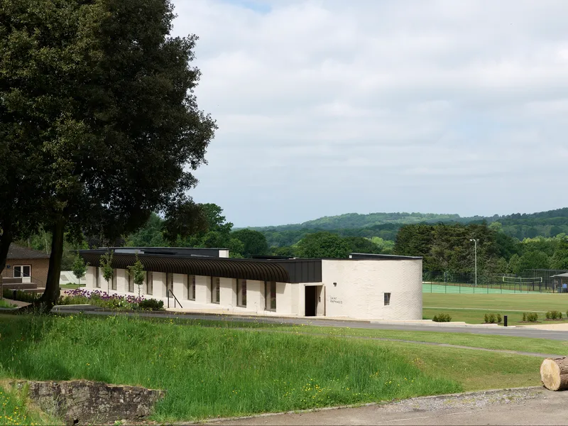Low-rise pavilion with corrugated barrel-vault roof and pale brick walls set beside a sports field