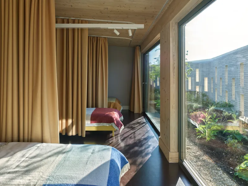 Bedroom with full-height glazing overlooking a planted courtyard framed by timber beams and ochre curtains