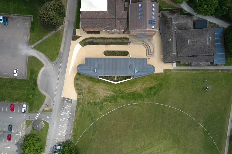 Aerial view of the curved pavilion with dark metal roof surrounded by gravel courtyard and grass