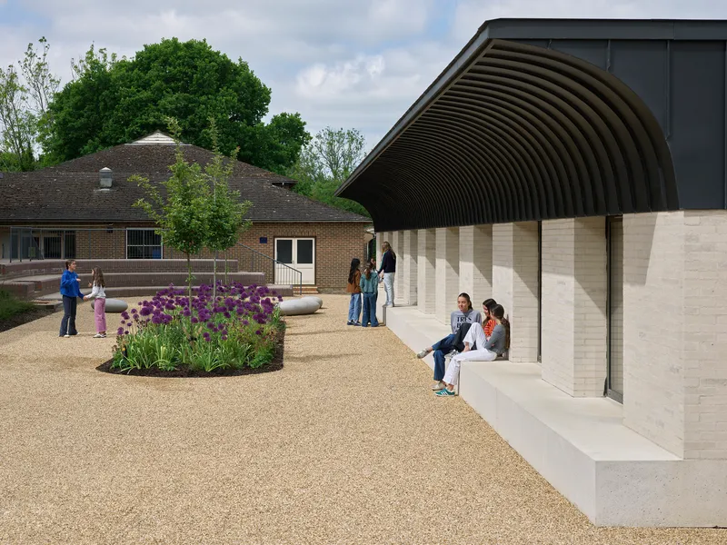 Courtyard with gravel surface and planted beds beside the ribbed metal canopy where visitors gather