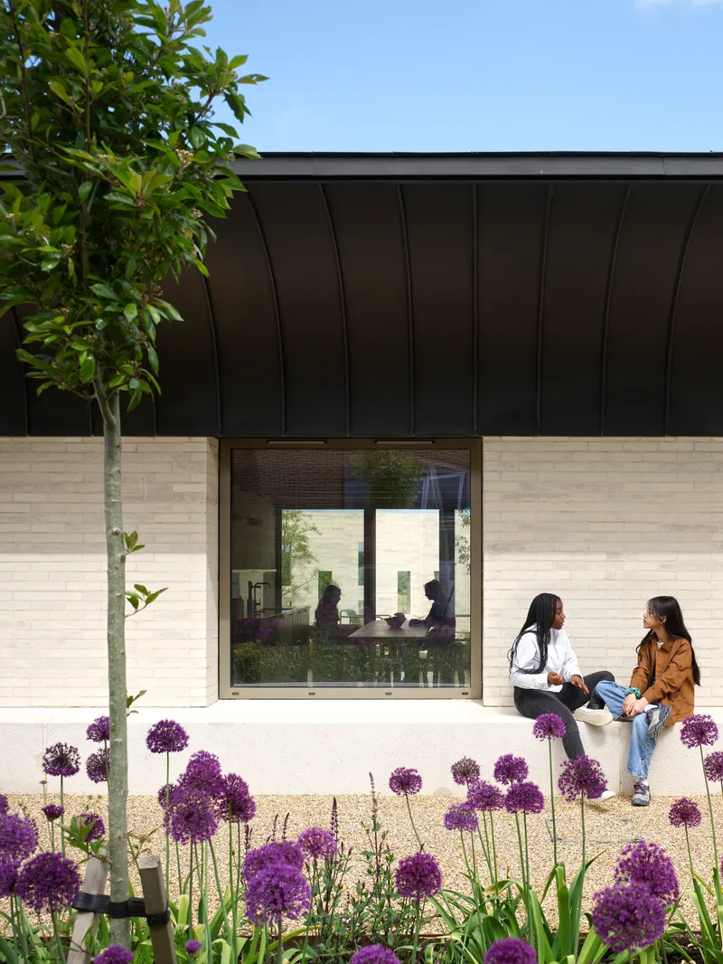 Cream brick facade with dark metal canopy and glass window framed by purple allium flowers