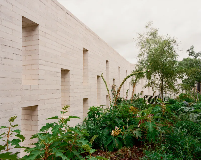 Pale brick wall with recessed vertical openings behind dense planting under an overcast sky
