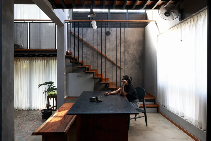 Double-height dining area with black metal staircase, timber table and bench, and sheer curtains in afternoon light