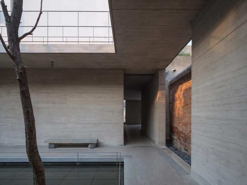 Courtyard view showing board-formed concrete walls, a tree trunk, and exposed brick remnant wall in afternoon light