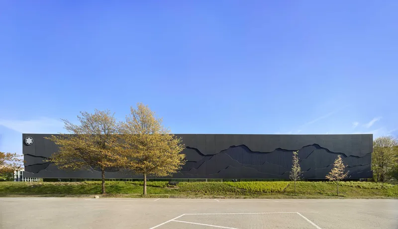 Dark metal facade with undulating relief pattern set behind a hedge and autumn trees