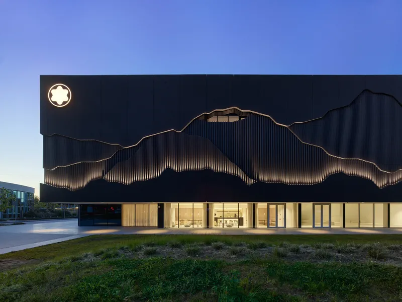 Illuminated glass-walled entry beneath dark metal cladding with wave pattern at dusk