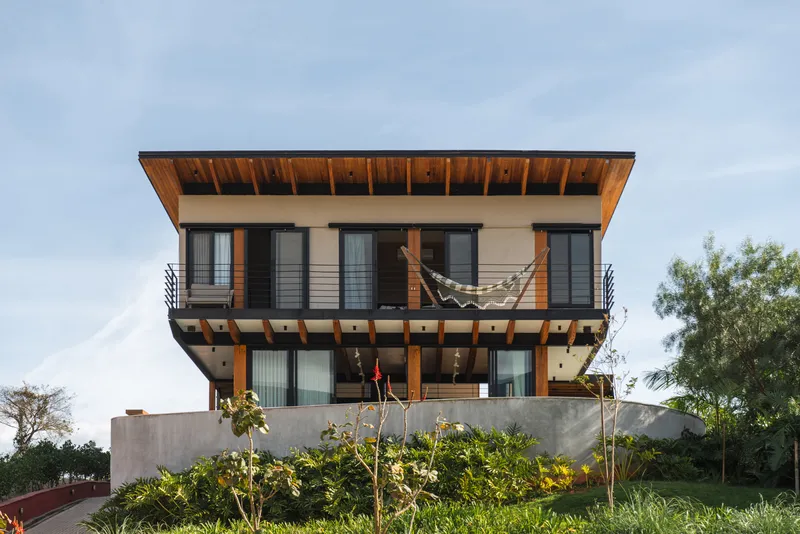 Front facade with exposed timber beams and overhanging roof above a concrete retaining wall and planted slope