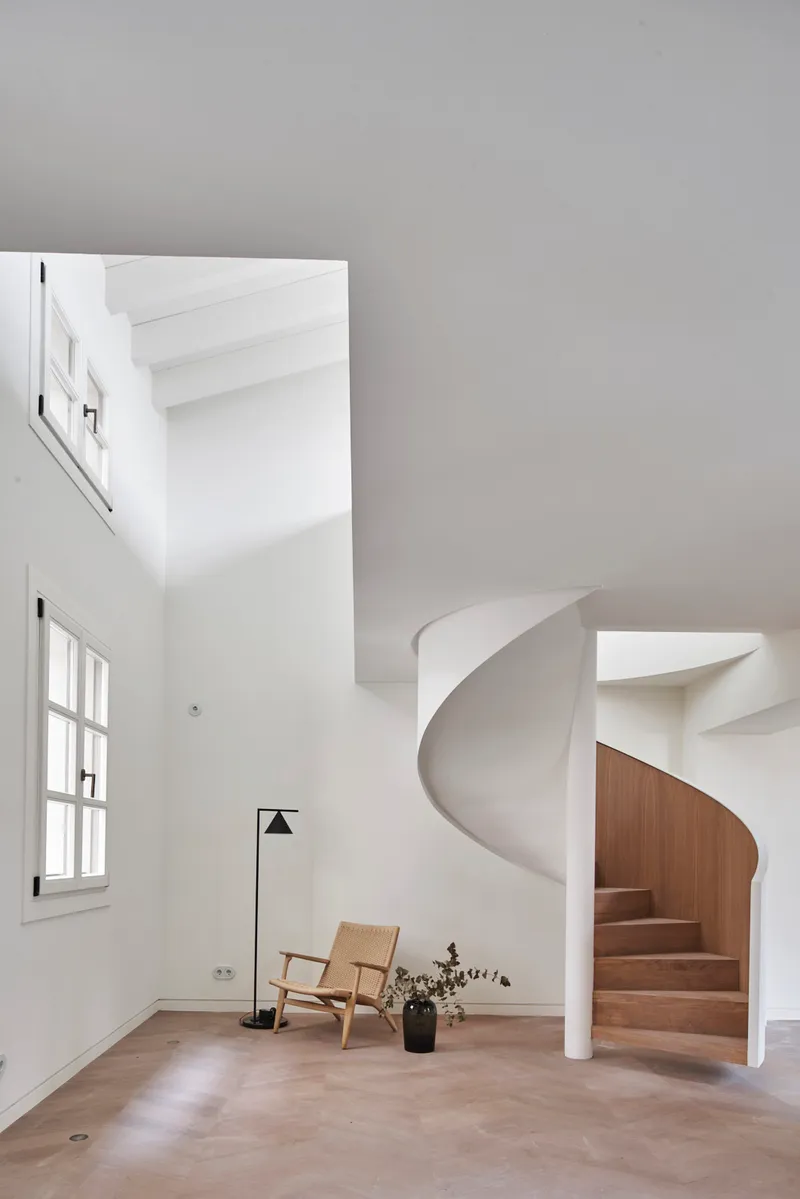 Helical staircase with white plaster volume and timber treads beside a canvas chair and floor lamp