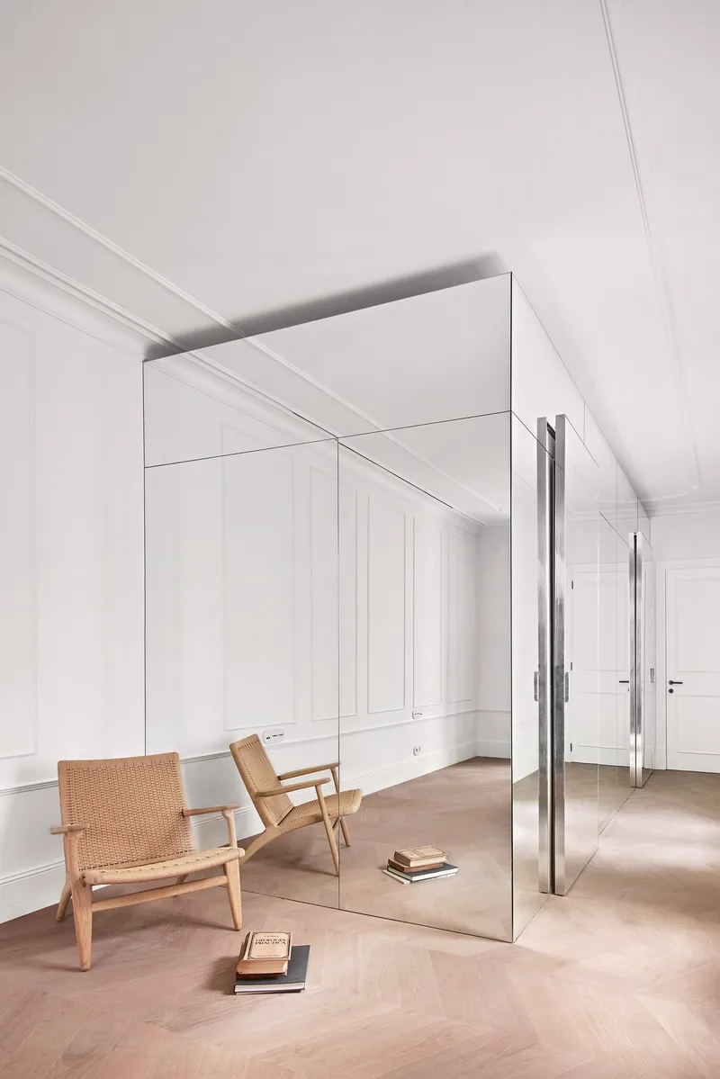 Freestanding mirrored volume with white paneled walls and herringbone oak flooring beside a woven chair