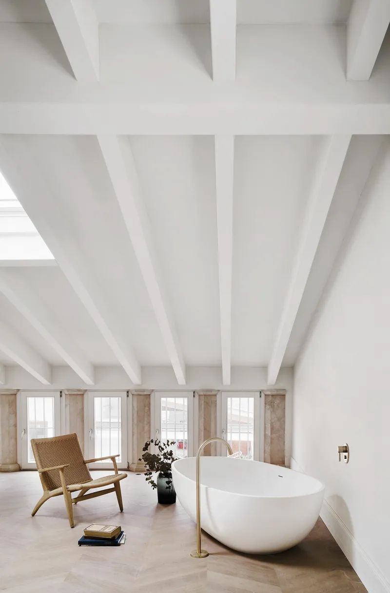 Bathroom with white painted beams and freestanding oval tub beside windows overlooking adjacent rooftops