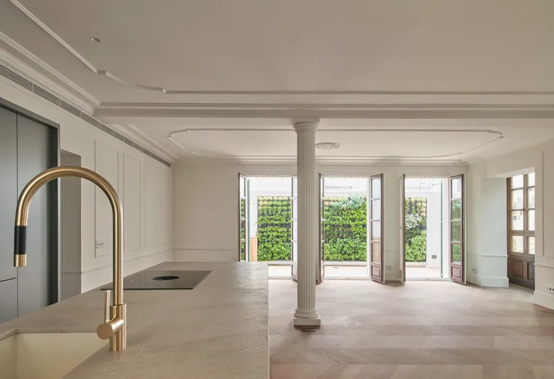 Kitchen island with brass faucet facing fluted column and tall windows opening to greenery outside
