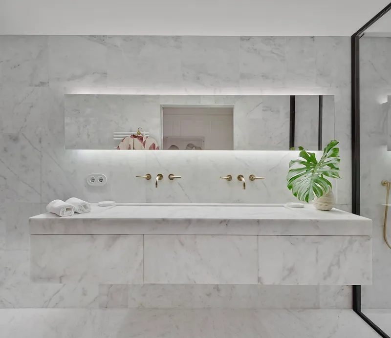 White marble vanity with integrated trough sink and brass wall-mounted faucets beneath a backlit mirror