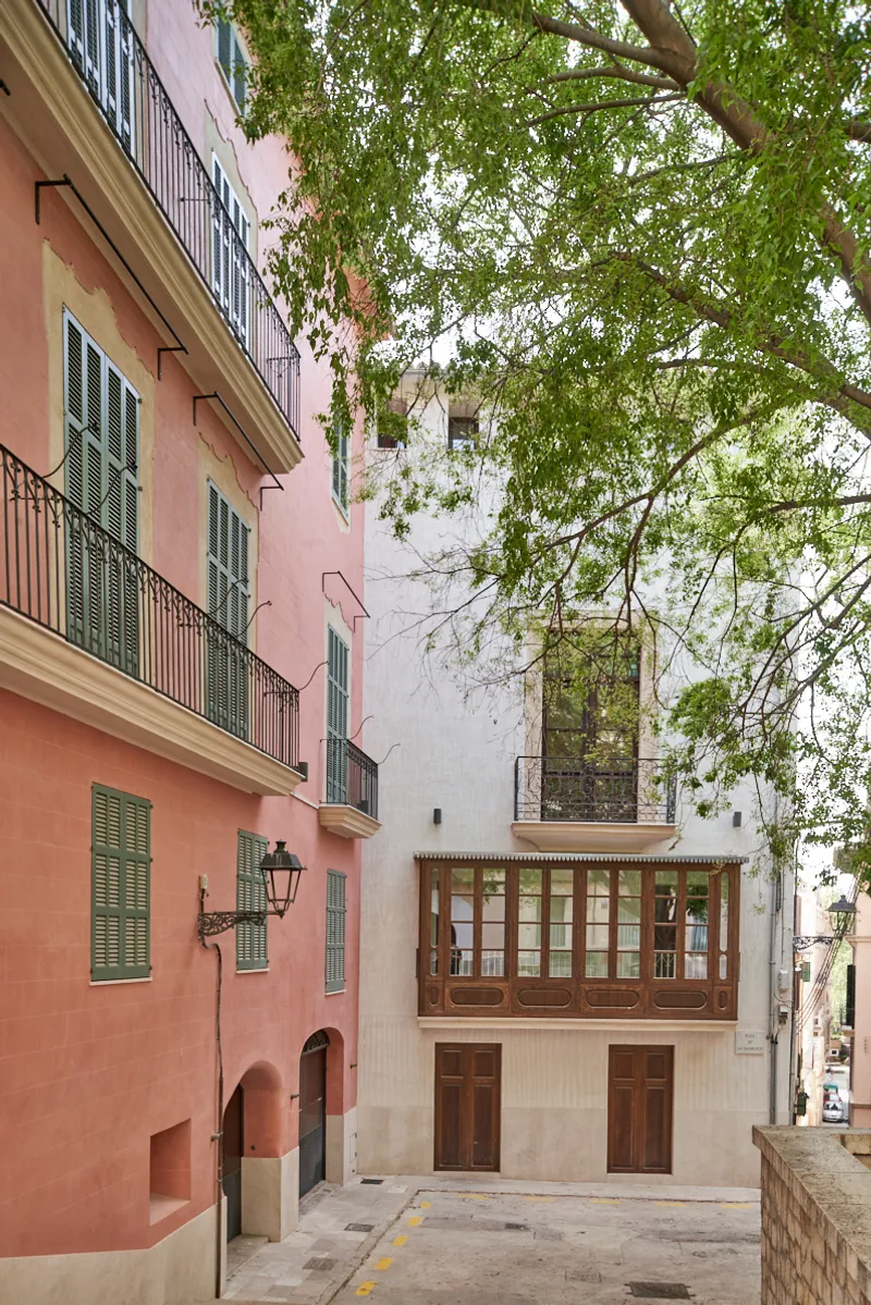 Narrow street view showing a pale facade with timber bay window between a coral building and trees