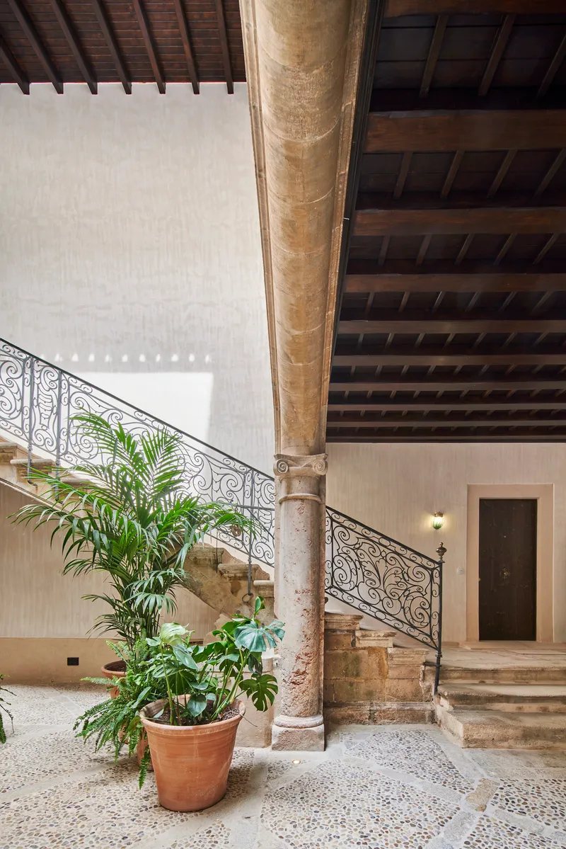 Stone staircase with wrought iron railing beside a weathered column and potted palm in a terrazzo courtyard