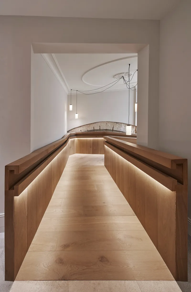 Central aisle between timber benches with glowing backlighting and suspended pendant lamps under curved plaster ceiling