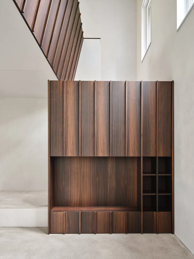 Freestanding walnut storage unit with vertical slat doors beneath a timber staircase in a double-height space
