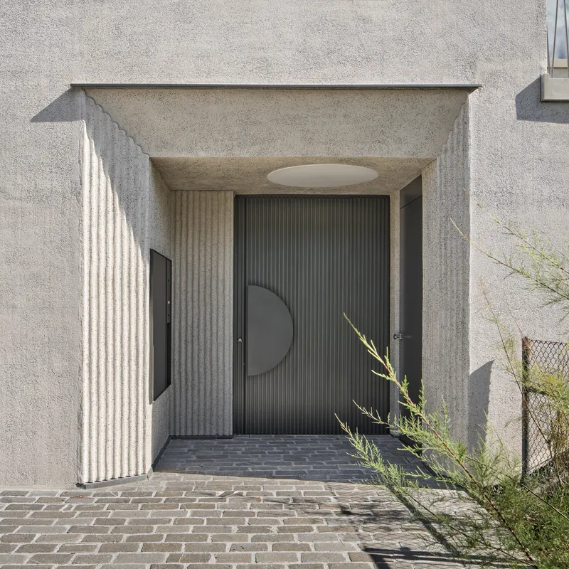 Recessed entry alcove with fluted concrete walls and dark ribbed door featuring a circular metal panel in sunlight