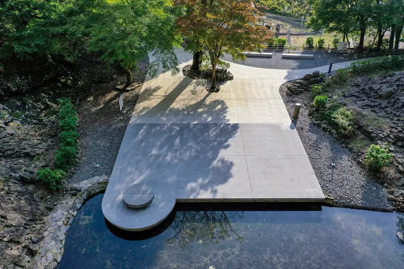 Concrete pathway extending over a reflecting pool with dappled tree shadows and a cylindrical stone element