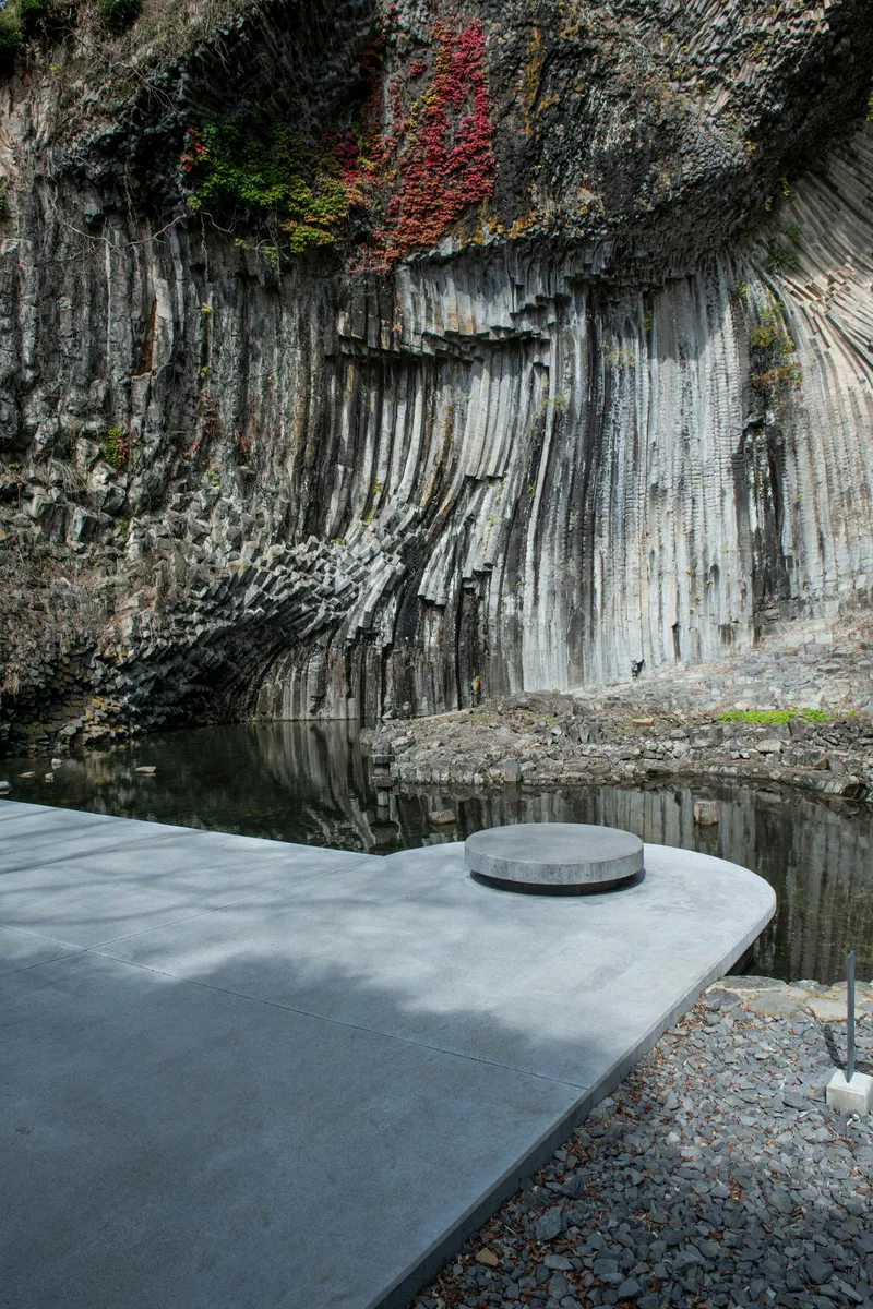 Curved concrete terrace with circular vent beside a still pool reflecting the columnar basalt cliff