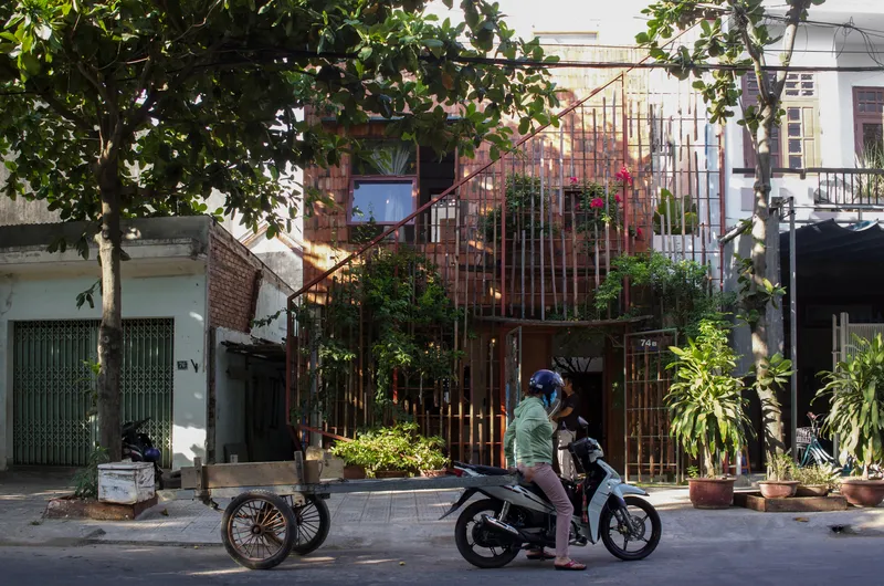 Street view of the vertical metal rod screen facade framed by overhanging trees and a passing motorbike