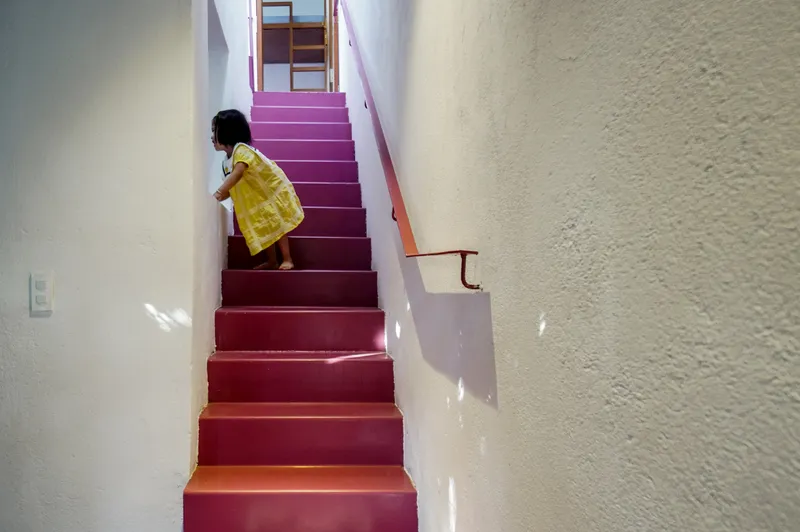 Child in a yellow dress ascending the maroon-painted staircase between white textured walls