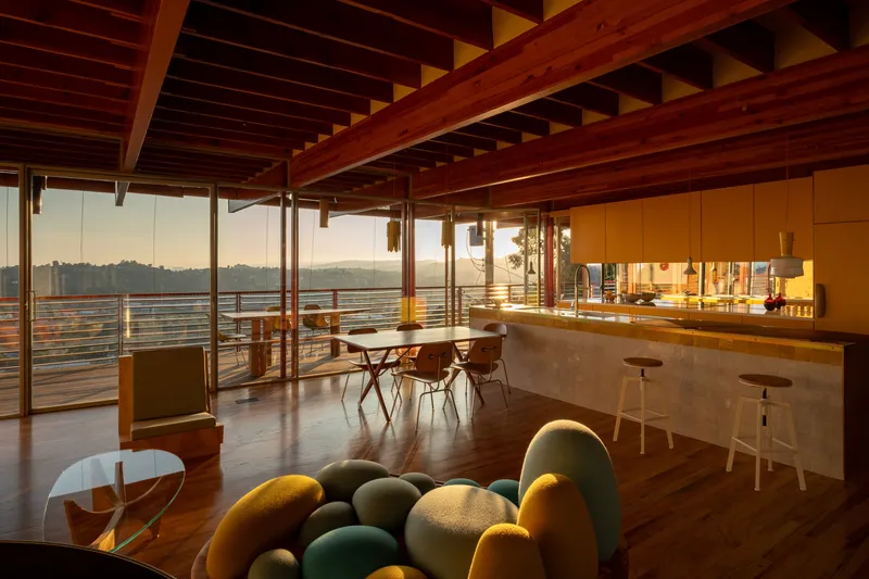 Open-plan living space with exposed timber beam ceiling and glass walls facing valley views at sunset